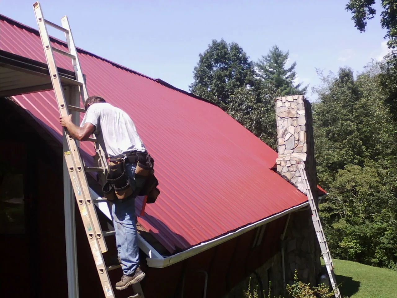 Roofer on a ladder beside a red metal roof and stone chimney — Soffit & Fascia Repair by Roofing Brothers in Arvonia